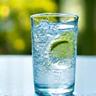 Bicchiere d'acqua cristallina con cubetti di ghiaccio e una fetta di limone, su uno sfondo luminoso.